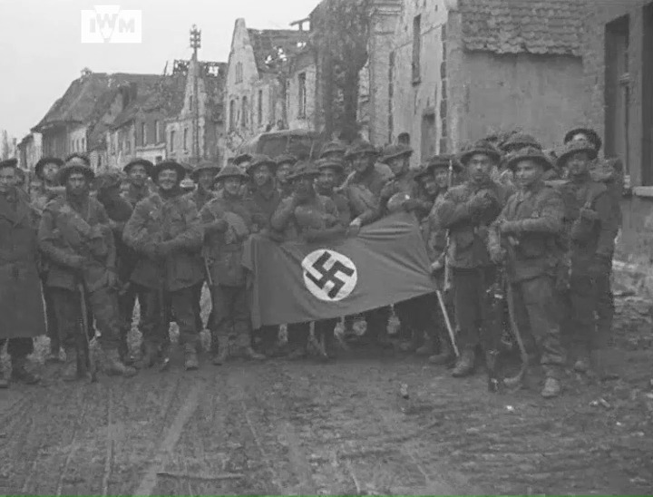 Men of the 10th Battalion, Highland Light Infantry, pose with a captured Nazi flag and sign reading Adolf Hitler-Strasse in Kranenburg, Germany, February 1945. Film: IWM A70 239-2
