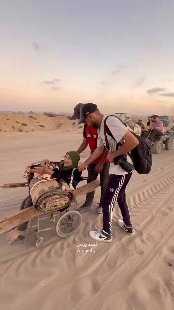 On her wheelchair and barefoot, the displaced elderly woman carries her mat and the weight of memories, while young people extend a helping hand along the way.