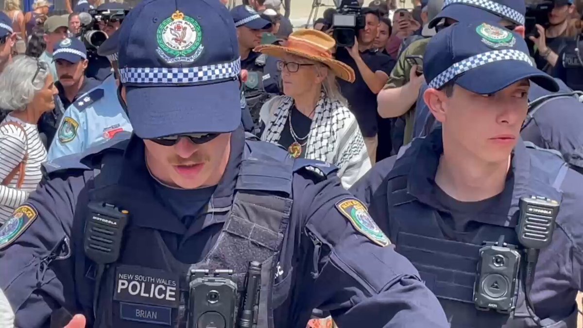 A woman in a kaffiyeh has been escorted by police away from the Bondi Pavilion site that has become a makeshift memorial to the 15 people murdered by terrorists yesterday.