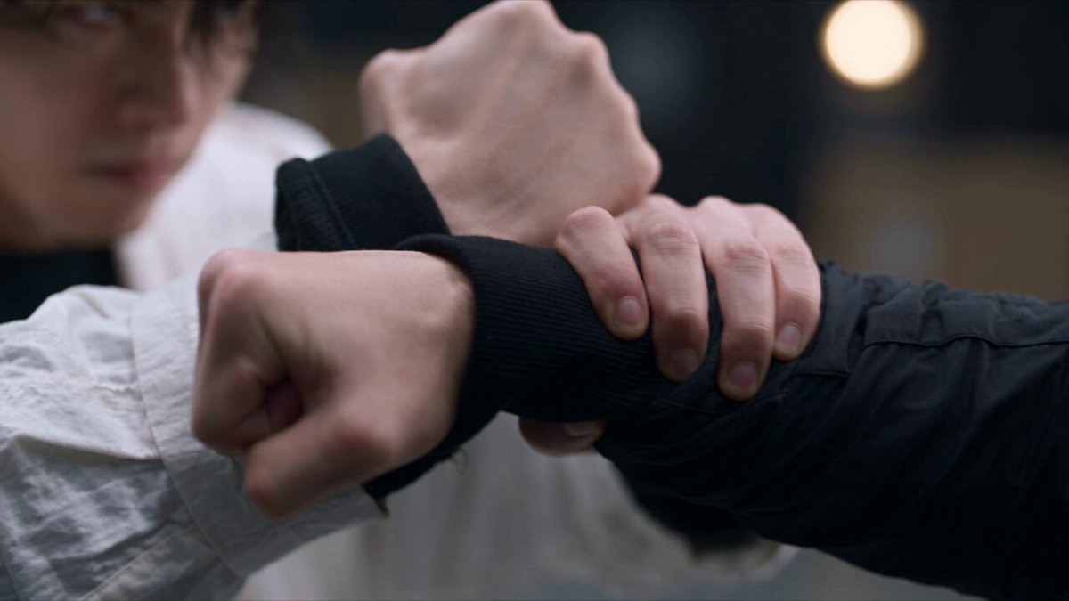 主提示词： 19秒，16:9，30fps，4K，写实电影级亚洲近身动作片，视觉上连续的一镜到底，专业武术指导，克制的少量血沫与口水，不出现重度血腥。 场景是一座宽阔的工业摄影棚兼仓库：灰蓝色水泥地面、红色定位线、黑色钢结构墙壁、黄色金属栏杆、堆叠纸箱和高处工业灯。冷灰色电影调色，局部暖黄色实景光，高反差照明，空气中存在轻微尘埃，背景保持真实而不过度虚化。 两名年轻亚洲男性进行无武器近身格斗。主角黑色中短发，穿飘逸的白色长款连帽外套、黑色内搭、黑裤和白鞋；对手身材高瘦、黑色短发，穿全黑连帽夹克、黑裤和黑鞋。两人的脸部、发型、服装、身高比例和左右位置全程一致。 0–2.1秒： 从两人拳头和手腕相互抵住的极近特写开始，画面中主要是交错的拳头、指节以及黑白袖口。浅景深，背景仓库化为柔和光斑。镜头沿两人的手臂缓慢横移并轻轻拉远，焦点从指节转移到白衣主角紧绷的眼神和面部。双方保持静态角力，手臂肌肉轻微颤动，营造爆发前的压迫感。 2.1–5.4秒： 动作突然加速。白衣主角踏步贴身突进，连续打出快速直拳、短摆拳和近距离前臂攻击；黑衣对手侧闪并抬臂封挡，随即挥拳反击。白衣主角以咏春式黏手连续截击，拍开手腕、改变攻击线路，低头钻入对方防线，压低重心打出短距离躯干拳。 镜头采用24毫米广角贴身跟拍，迅速从手部特写拉到双人中景，随后贴着肩膀顺时针环绕。手臂数次掠过镜头形成自然前景遮挡，伴随快速甩镜、轻微荷兰角和真实运动模糊。镜头运动激烈但始终能够辨认双方位置和动作方向。 5.4–6.3秒： 白衣主角从低位打出向上的掌根或前臂攻击，准确命中黑衣对手下颌。镜头从胸口高度快速下沉并推到对手面部的极低机位特写。命中瞬间，黑衣对手面部产生短暂的真实挤压，头部沿受力方向向侧后方甩动。 对手嘴角喷出少量半透明口水细雾，混合三到五颗暗红色小血沫。液滴严格沿打击方向飞散，在工业逆光中短暂闪亮，随后受到重力影响落下。下唇和嘴角留下非常轻微的血迹，没有开放性伤口。使用短暂速度坡度慢动作突出击中、液滴飞散和痛苦表情。 6.3–9.5秒： 速度恢复。黑衣对手横向挥臂反击，白衣主角双手交叉截住攻击，控制手腕并向下牵引，低头从手臂下方钻过，绕到对手侧面。双方进行连续的手臂缠斗、反抓、格挡、肘部封锁、肩部撞击和近距离换位。 白衣主角拍开对手拳头，以开放手掌托起下颌，同时控制另一只手腕；黑衣对手扭转身体尝试挣脱。镜头从两人之间穿行，紧贴手掌、手腕和面部进行横向弧形运动。衣袖偶尔遮住大半画面，遮挡结束后自然显露新的攻防位置，不出现硬切。 9.5–10.2秒： 白衣主角移动到黑衣对手侧后方，一只手控制手腕，另一只前臂压住颈侧，使对方失去平衡。镜头形成倾斜的面部近景，黑衣对手咬紧牙关并挣扎，嘴角仍保留很少的血迹。动作短暂放慢，强调颈部控制和身体重量。 10.2–11.6秒： 镜头顺着两人的身体快速下沉至贴近地面的低机位。白衣主角稳定支撑脚步，以膝击接低位侧踢破坏对手重心。白色长外套从镜头前方掠过，形成强烈的白色动态前景。 在腿部完全伸展、对手身体开始倾斜的瞬间进行约一秒的速度坡度停顿。能够清楚看见鞋底、裤脚、重心转移和衣料惯性，随后恢复正常速度。 11.6–13.3秒： 黑衣对手弯腰后强行旋转身体，尝试以反身肘击和横扫摆拳反击。白衣主角后撤半步避开肘尖，随即贴身进入内侧，截住前臂、转动髋部并把攻击引向一旁。 镜头跟随两人重新升高，从低机位迅速抬升至胸口高度，并以弧形轨迹环绕。高速阶段产生自然背景拖影，仓库中的栏杆、纸箱和地面红线形成明显的空间参照，最后拉到能够看清两人全身关系的中广景。 13.3–14.4秒： 两人在极近距离同时停住，拳头分别悬在对方面前，身体保持蓄力状态。白衣主角略微侧身，黑衣对手后脚支撑、上身前倾。动作近乎定格，只有呼吸、眼神、衣摆和肌肉存在细小运动。 镜头由激烈手持突然变为稳定的缓慢横移，形成短暂而压迫的安静，让观众清楚看见双方距离和下一击路线。 14.4–16.3秒： 白衣主角突然爆发，拍开对手的防守手臂，踏入内线，以短距离肘击或前臂横击重重命中黑衣对手的下颌与颈侧。动作短促、准确、有明显的髋部发力和肩膀传导。 镜头高速甩入极近面部特写，在接触瞬间产生真实横向运动模糊和一次克制的冲击震动。黑衣对手的脸沿受力方向转动，一小团扇形口水雾从嘴边喷出，其中混合少量暗红血点。血量非常克制，只出现几颗飞散血滴和嘴角血迹，没有大量喷血、伤口或牙齿脱落。 命中后进入约一秒半的速度坡度慢动作。黑衣对手眼神失焦，头部自然歪落在白衣主角的前臂上；细小液滴继续沿原方向飞行并迅速落下，白色衣袖只沾上一两颗很小的暗红血点。 16.3–19.1秒： 白衣主角旋转撤步并释放对手。黑衣对手失去支撑，向后倒地、翻滚一次，最终趴倒在远处地面。身体落地具有真实重量和惯性，不弹跳、不漂浮。 镜头随着白衣主角的旋转迅速拉到低机位中广景。白衣主角背对镜头站在画面前景，长款白色外套因旋转大幅展开，在空气中形成圆弧状飘动，并逐渐缓慢落下。黑衣对手位于背景左侧，先挣扎一下，随后完全倒下。 镜头最终稳定保持英雄式背影构图，轻微向前推进。白色衣摆占据视觉中心，仓库的巨大纵深完整展现，空气中的尘埃和几颗极小液滴在逆光中一闪而过。 19.1–19.3秒： 白衣主角保持背影站立，黑衣对手倒地不动，画面快速而平滑地淡出黑场。 整体视觉要求： 电影级真实人体动力学，专业近身格斗编排，每次攻击都包含预备、发力、接触、受力和回收。拳头、手掌与身体存在准确接触点，受击方向符合物理规律。衣服具有真实重量、褶皱和惯性，白色长外套形成富有表现力的动态轮廓。 模拟24毫米至28毫米广角电影镜头，贴身稳定器结合克制手持感，浅景深焦点转换，甩镜、弧形环绕、低机位、前景遮挡、轻微荷兰角、真实180度快门运动模糊。主要依靠镜头移动和遮挡完成转场，没有明显硬切。 口水呈半透明细雾，血液为少量暗红小血点，飞散方向必须与冲击方向一致，不悬浮、不突然出现、不持续喷射。整体具有强烈冲击力，但保持写实、克制，不走恐怖或重口味路线。 负面提示词： 大量喷血、血柱、血池、持续流血、整张脸被血覆盖、开放性伤口、断肢、内脏、骨骼外露、牙齿大量脱落、恐怖特写、夸张血浆、血液漂浮、口水过多、液滴逆着受力方向飞行、多余人物、多余手臂、多余手指、手指畸形、关节反折、肢体穿模、拳头穿过身体、没有接触就受伤、人物瞬移、滑步漂移、违反重力、角色换脸、发型变化、服装互换、身高变化、黑白衣服颜色混乱、左右位置突然交换、镜头轴线跳变、无意义剧烈抖动、全程慢动作、过度径向模糊、卡通特效、超能力光效、字幕、水印、Logo。