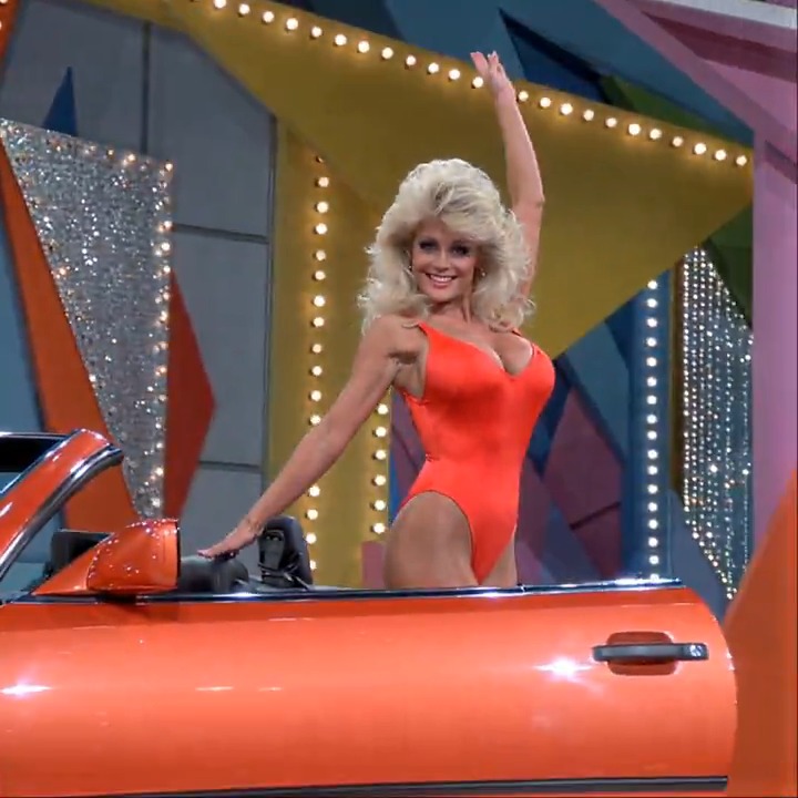 Blonde in a bright red swimsuit poses and waves next to a shiny orange Mercedes convertible on a colorful game show stage. Would you take the car or the moment? Credit awesome_visuals #PrizeReveal #RedSwimsuitPose #OrangeMercedesWin